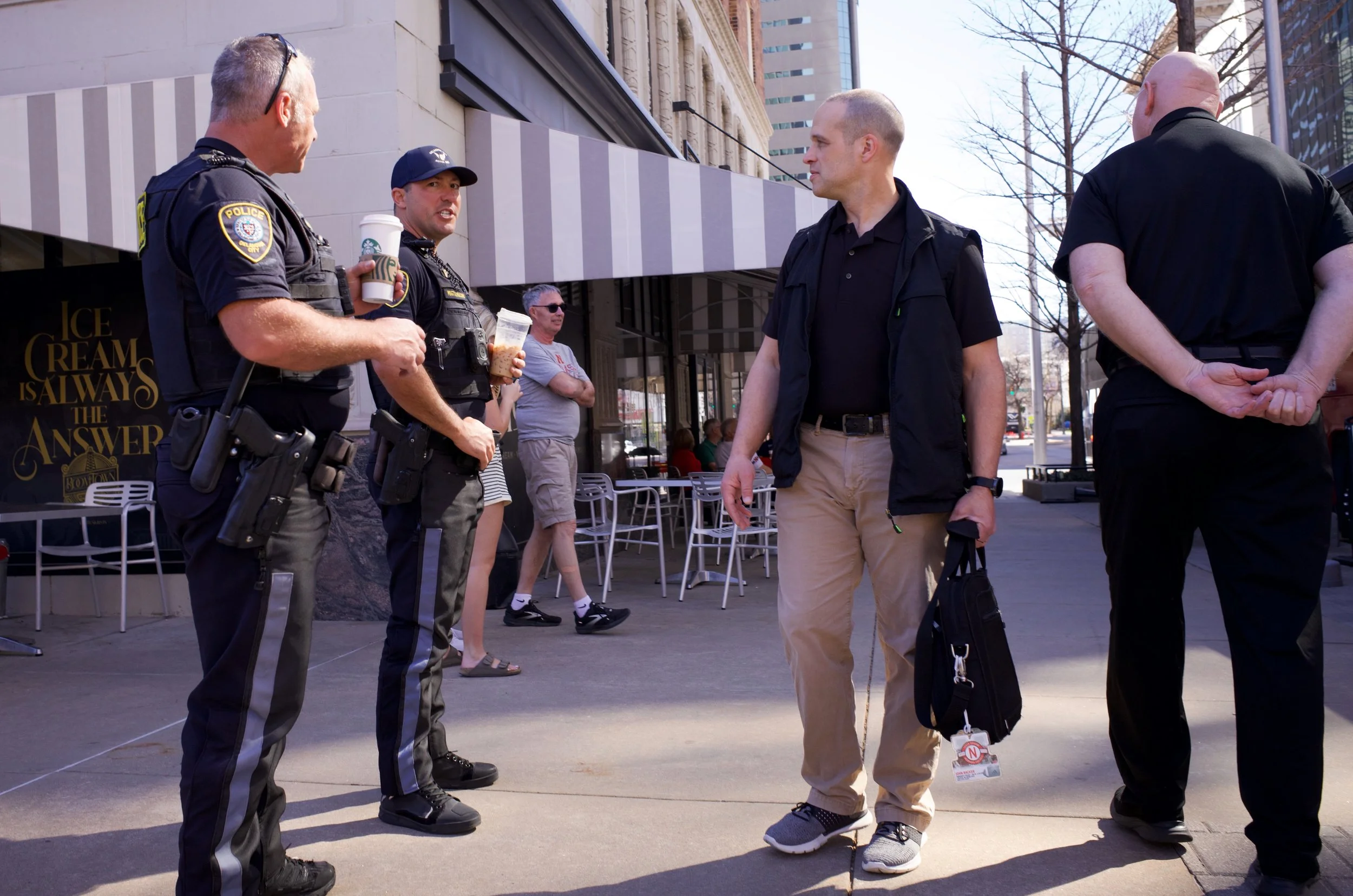 Four police officers and three civilians stand on a city sidewalk engaged in conversation. The police officers are in uniform, one holding a coffee cup and a bag, and they've got weapons on their belts. The civilians are casually dressed, with two pe
