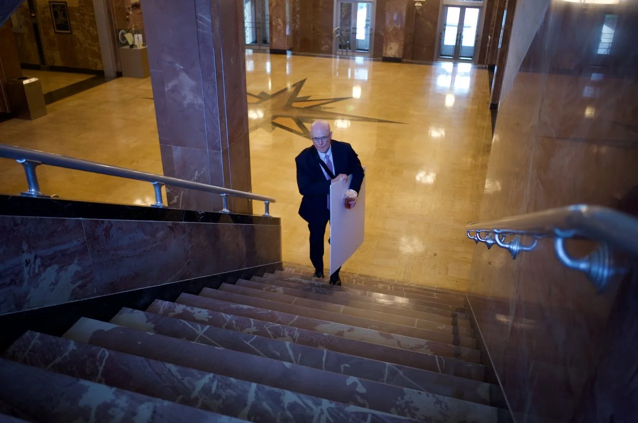 A man in a suit and glasses standing at the top of a staircase in a lobby, holding a drink and a sign, with an ornate clock and elevators in the background.