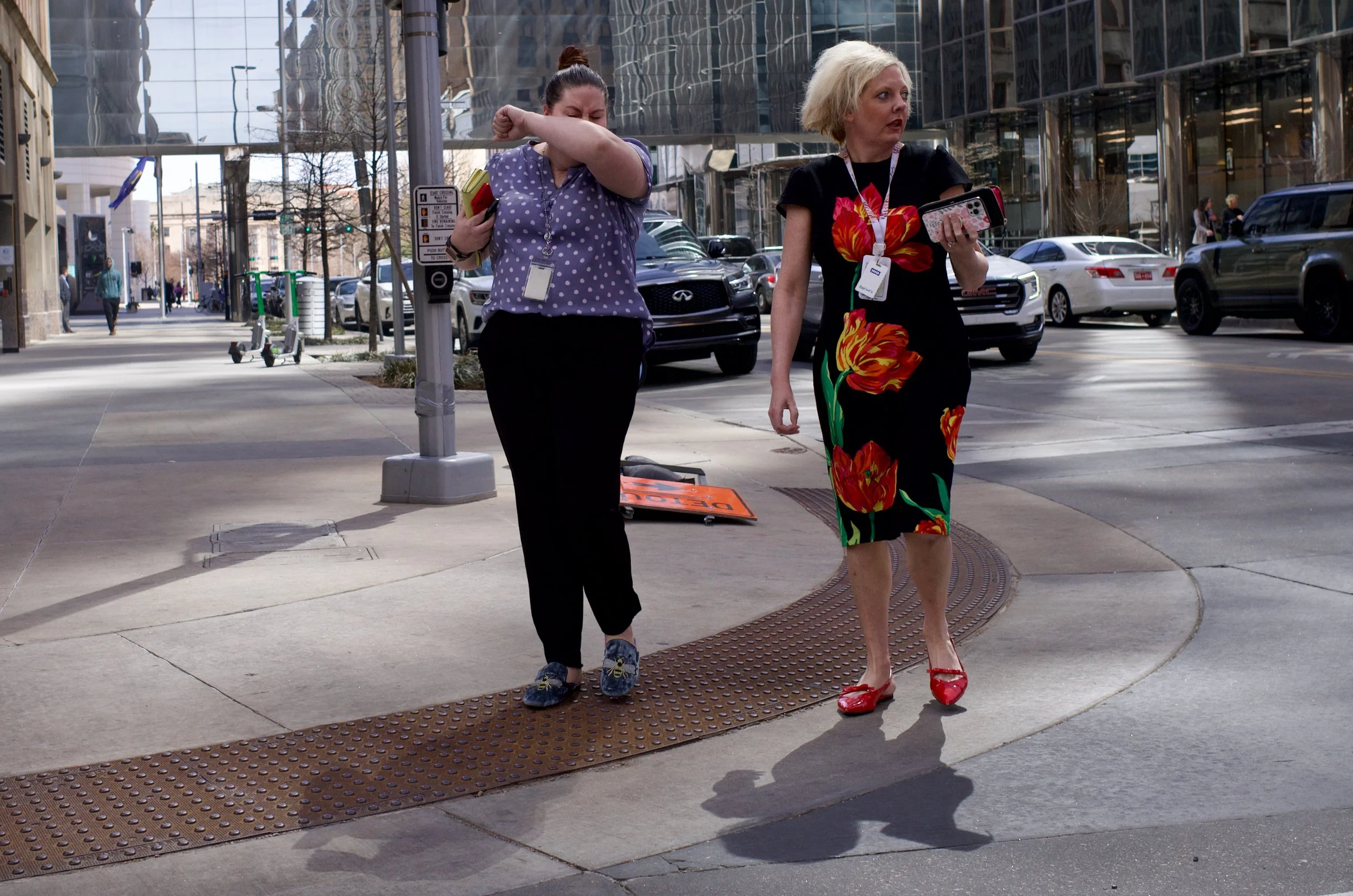 Two women walking on a city sidewalk, one is sneezing or wiping her face, the other is looking at her phone, both with event badges around their necks. The woman on the left wears a purple polka-dot shirt and black pants, and the woman on the right w