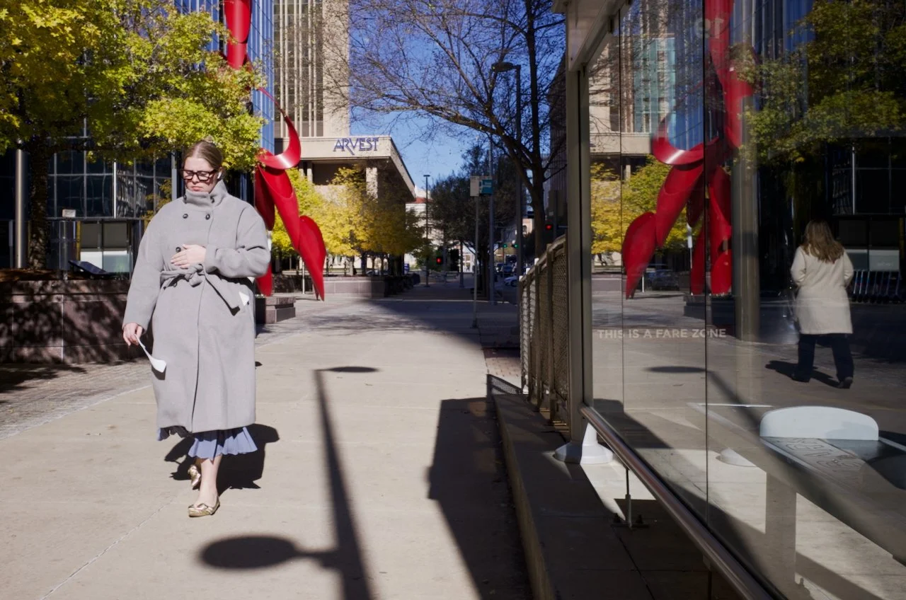 A woman in a gray coat and glasses walking on a city sidewalk, with trees, buildings, and reflections visible in the glass storefronts