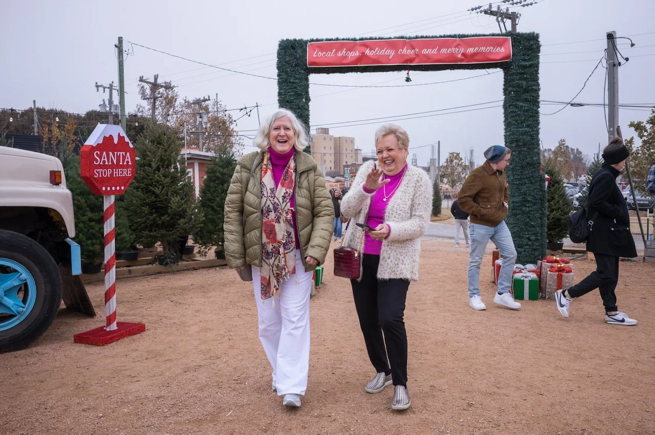 Two women smiling and walking in a Christmas tree lot decorated with holiday banners and signs, including a red sign that says "Santa Stop Here." Other people are in the background, and the area is outdoors with dirt ground and trees.