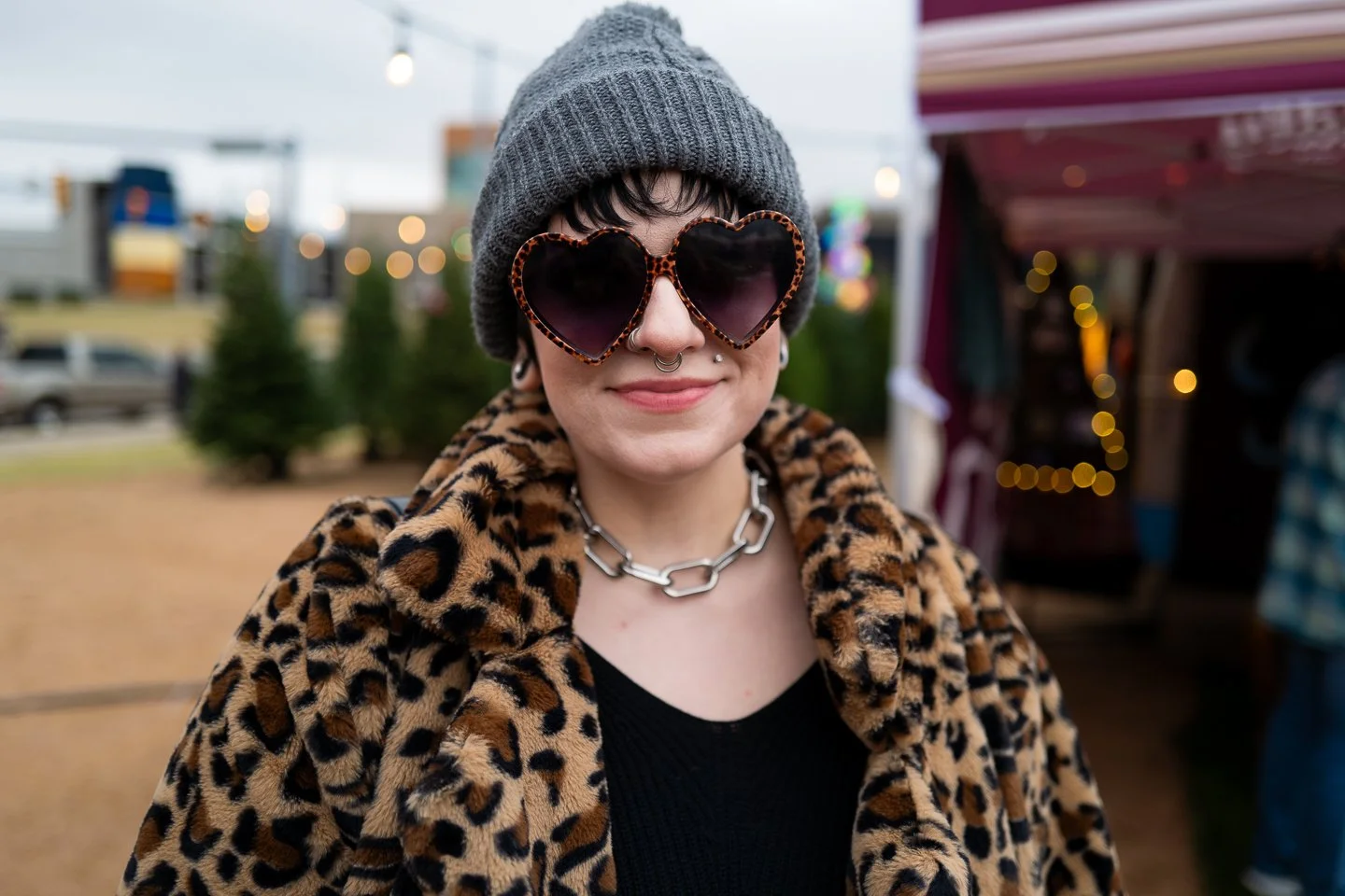 A woman in a gray beanie and leopard print coat with heart-shaped sunglasses, jewelry, and pink lipstick at an outdoor event.
