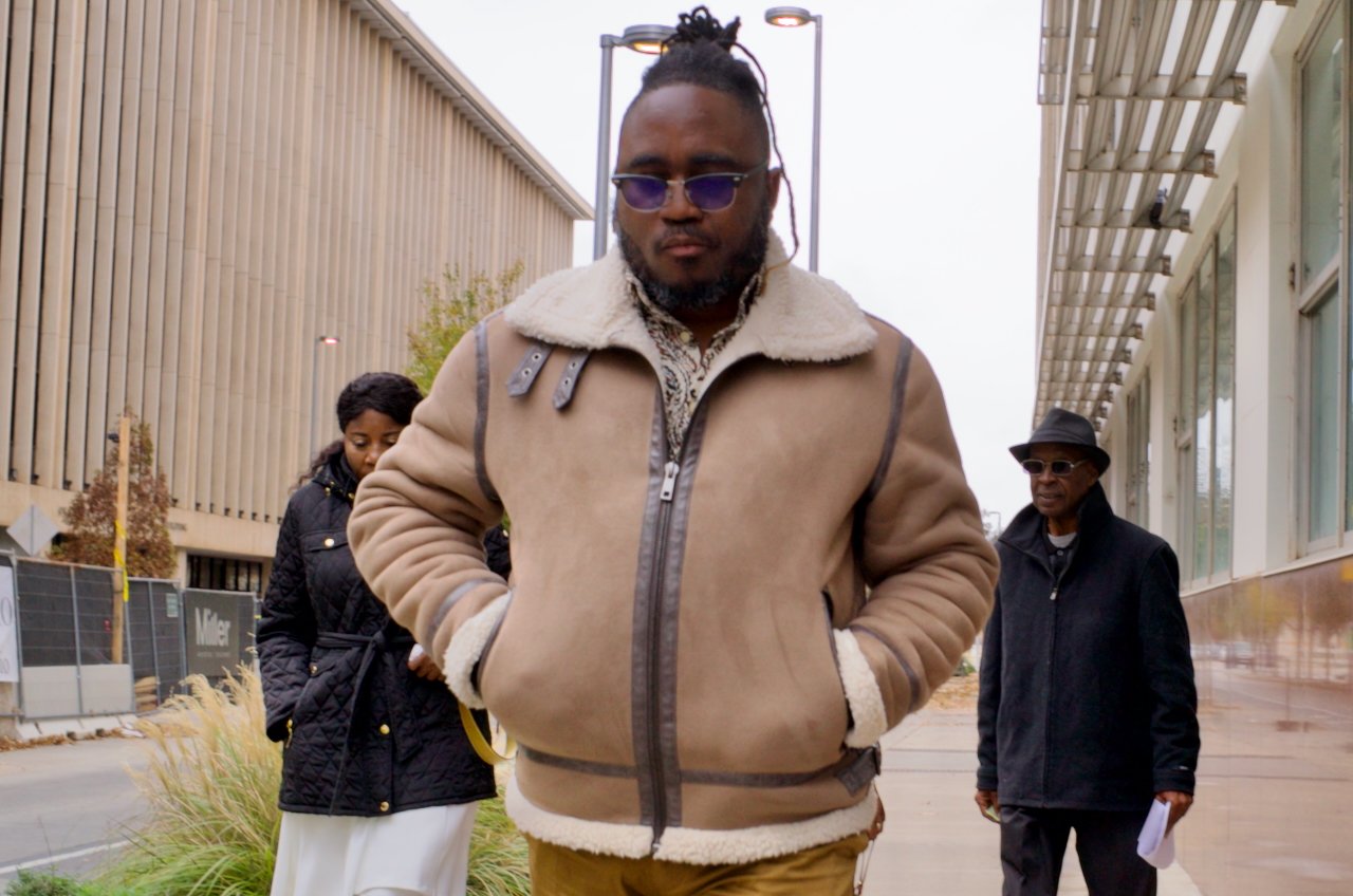 Man with sunglasses and a beige jacket walking outside with two people in the background during daytime.