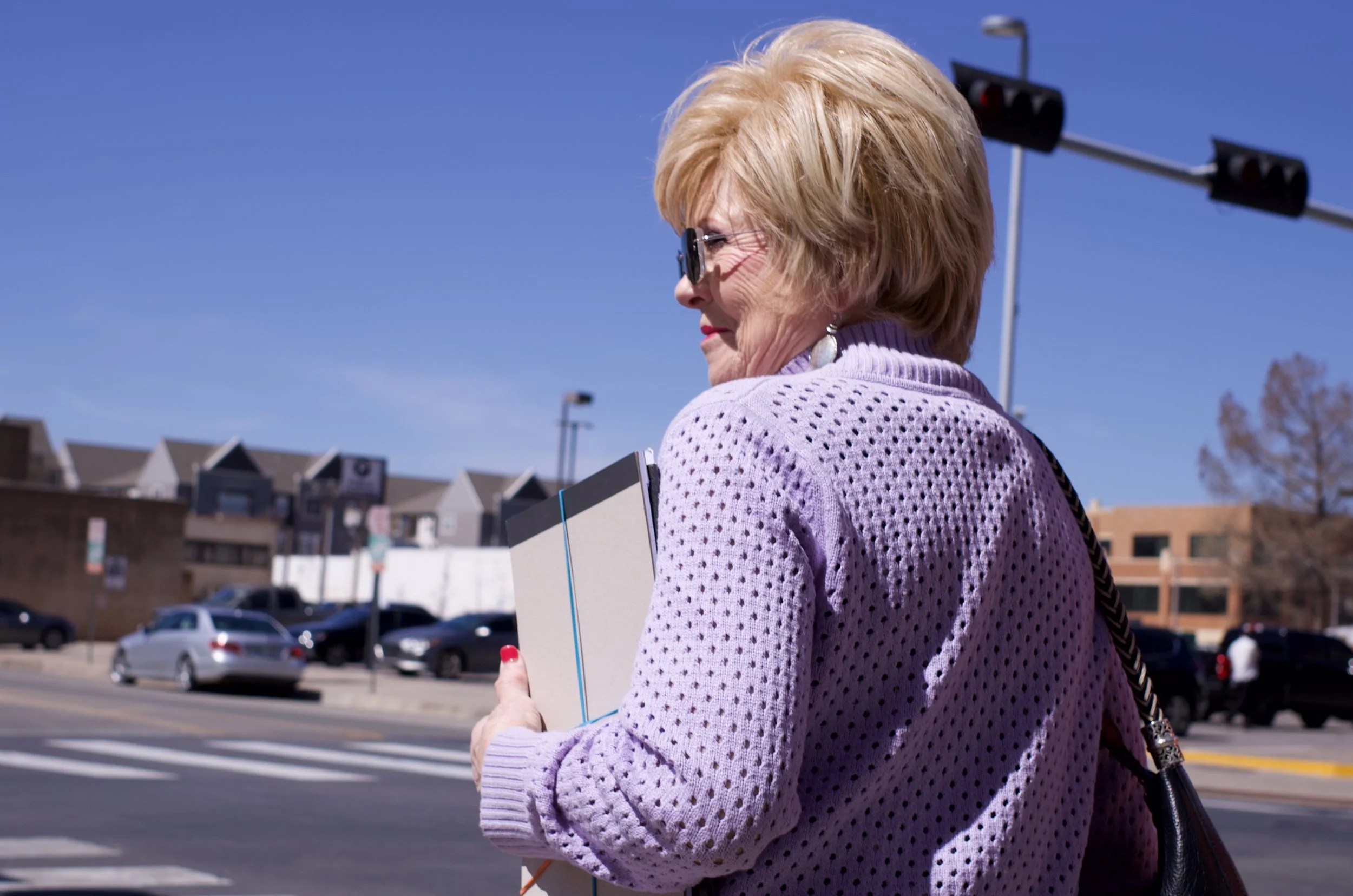 An elderly woman with blonde hair, sunglasses, and earrings, holding a notebook and a folder, walking on a crosswalk in a city with a traffic light overhead and parked cars in the background.