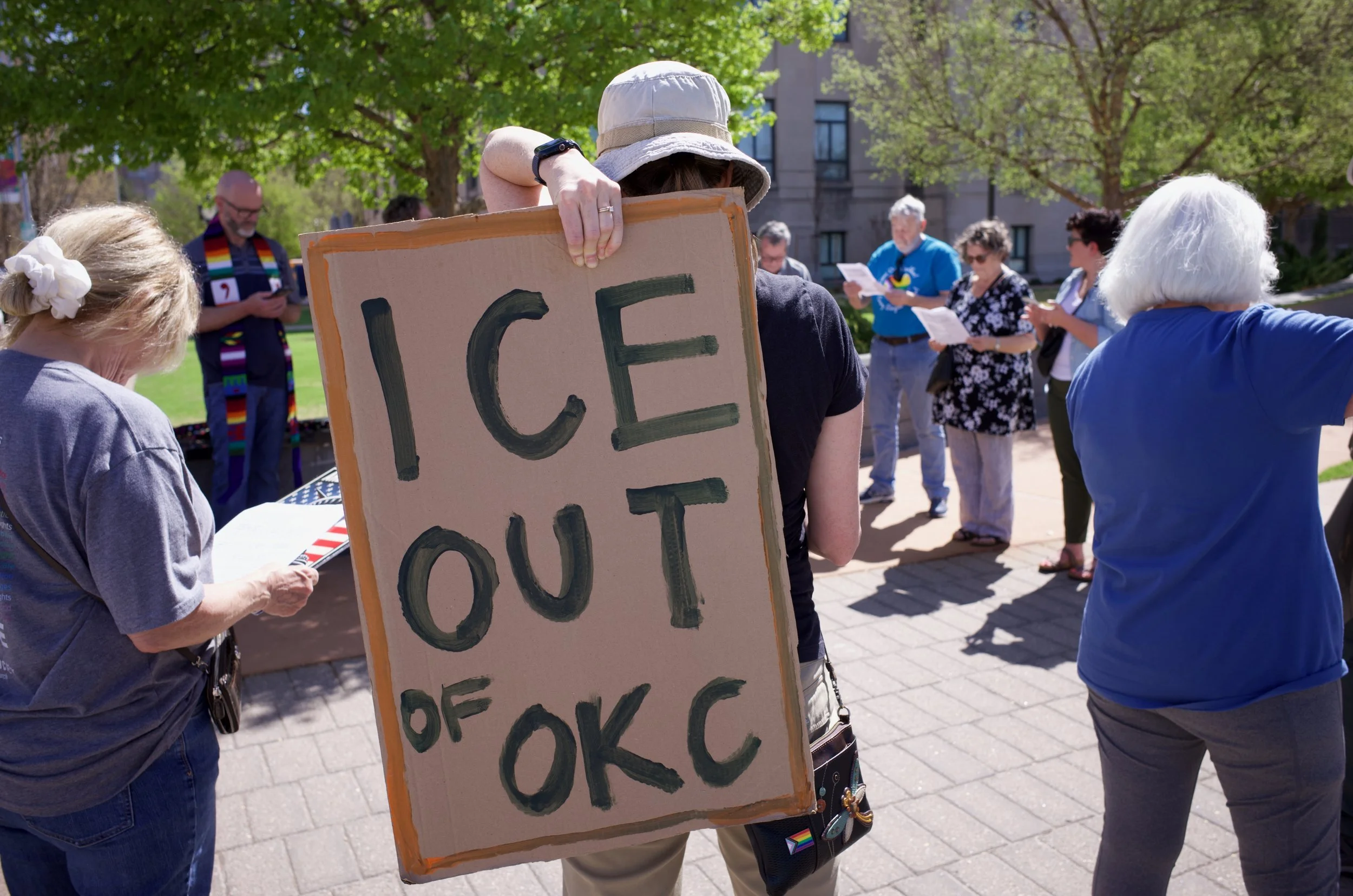 A protester holding a cardboard sign that says 'ICE OUT OF OKC' with several other people gathered in a park area.