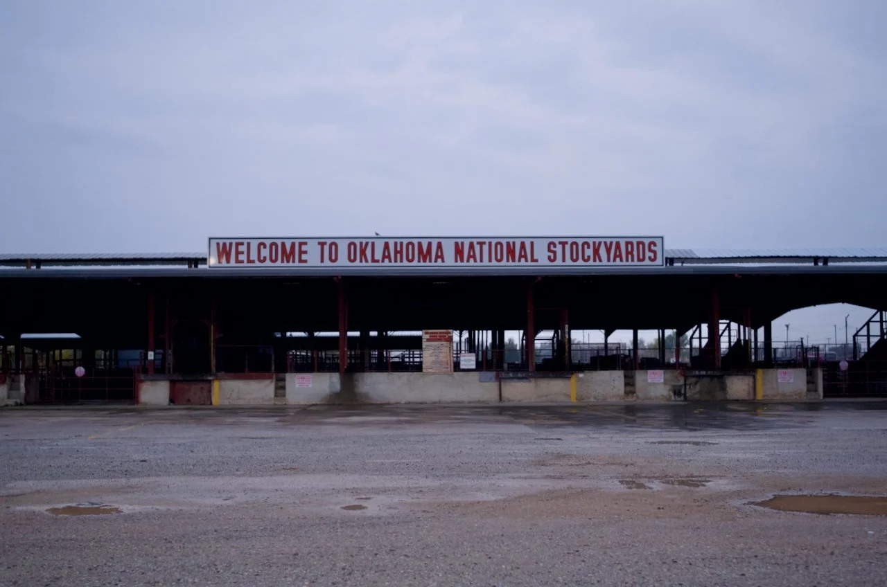 Entrance to Oklahoma National Stockyards with a large sign that reads 'WELCOME TO OKLAHOMA NATIONAL STOCKYARDS' under a cloudy sky.