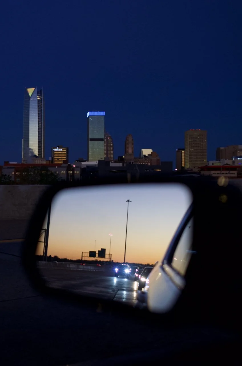 City skyline at dusk visible in rearview mirror on a highway, with cars driving and streetlights beginning to turn on.
