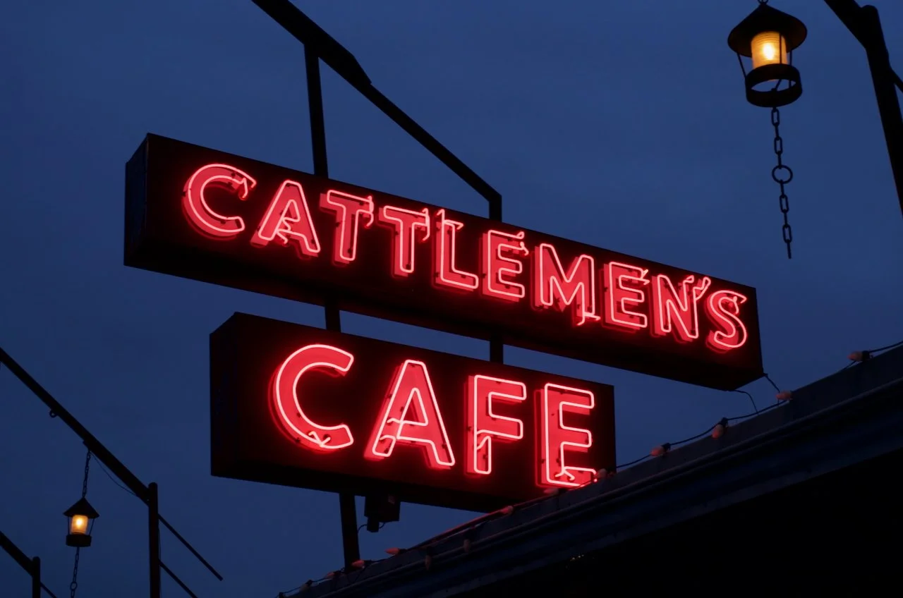 Neon sign reading 'Cattlemen's Cafe' against a blue evening sky with hanging lanterns.