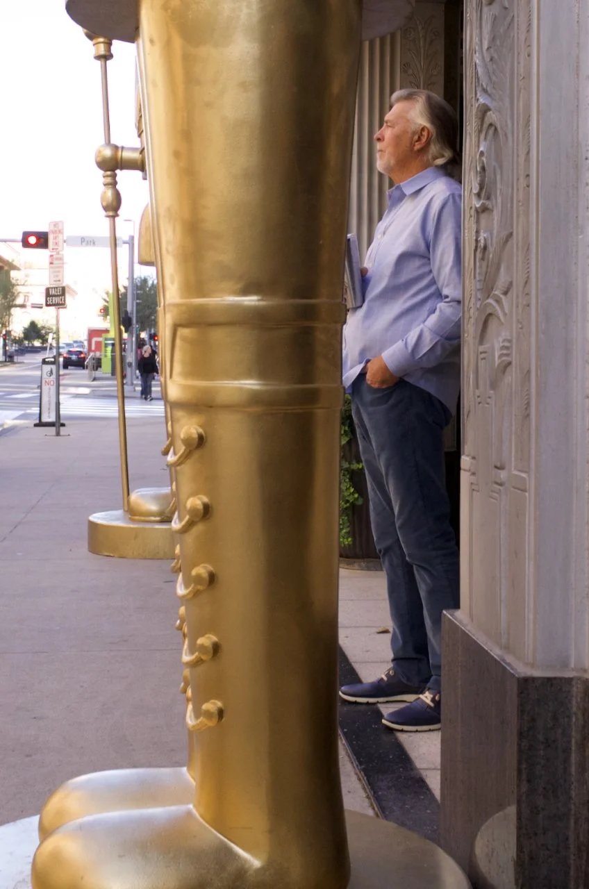 A man in a light purple shirt and dark pants standing outside, partly obscured by a large golden object, in an urban setting.