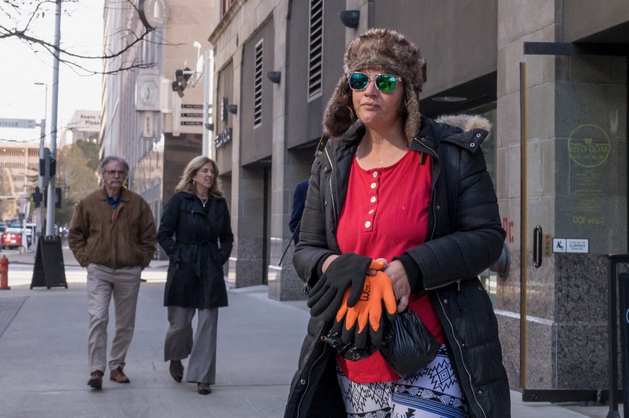 A woman in a red top, black coat with a fur-lined hood, wearing a fur hat and sunglasses, is holding orange gloves on a city sidewalk. Behind her are two older adults, a man and a woman, walking and talking. The man is wearing a brown jacket and beig