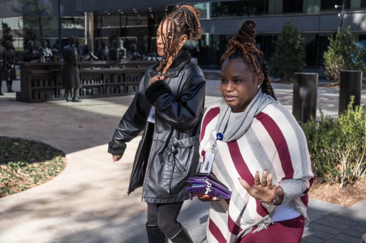Two women walking outdoors, one with a gray and maroon striped shawl, holding a purple folder, the other wearing a black leather jacket with braided hair
