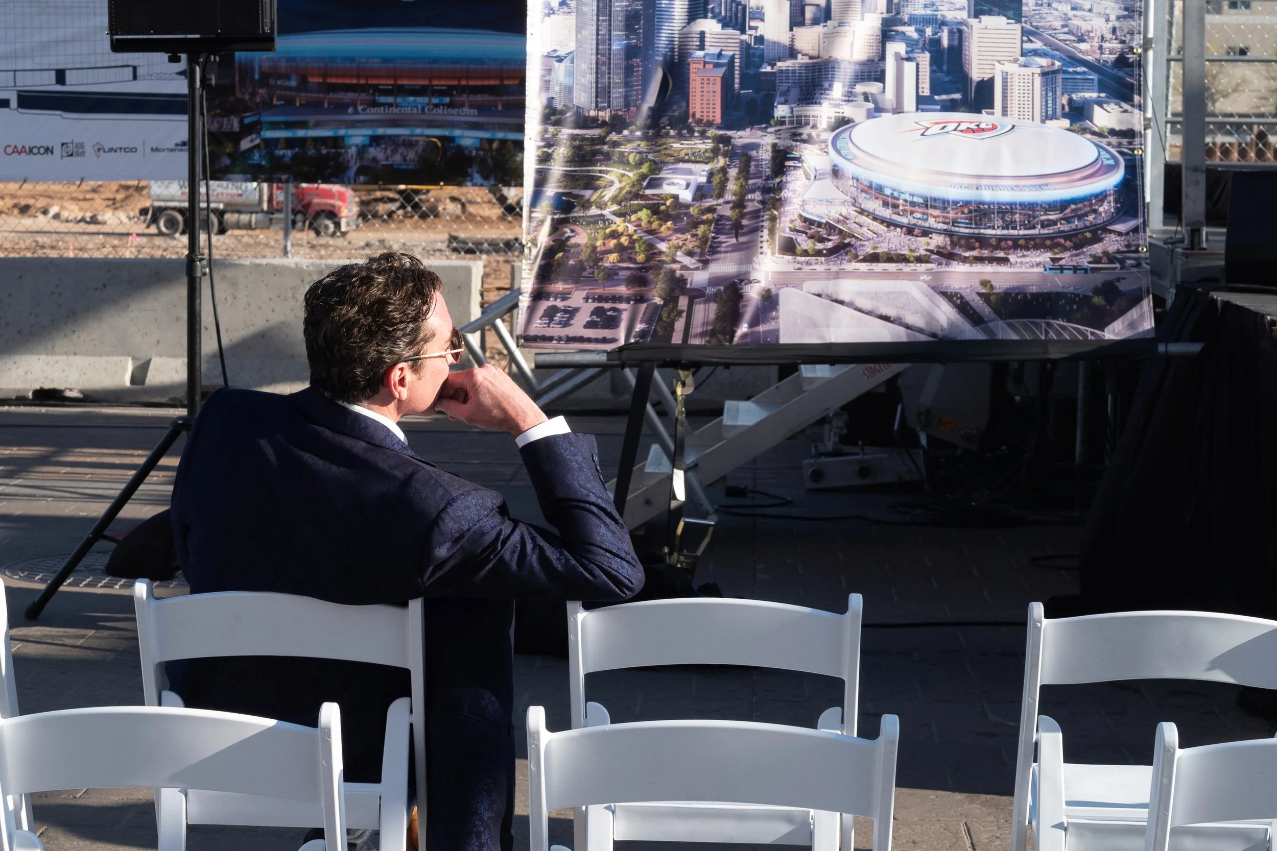 A man in a suit sitting on a white chair, looking at an architectural rendering of a stadium and surrounding city landscape on a display board at a construction site.