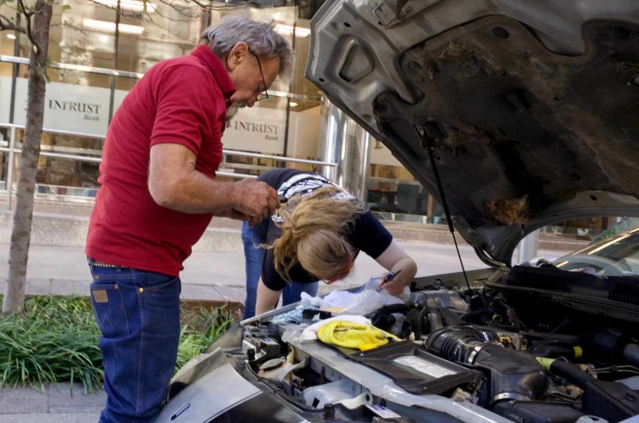 Two people working on a car engine with the hood open, outdoors on a city sidewalk.