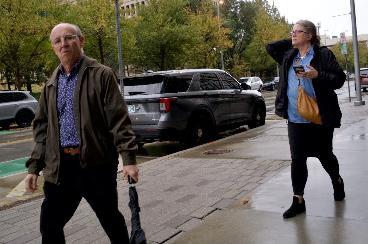 A man walking on a city sidewalk holding an umbrella, and a woman standing nearby looking at her phone on a rainy day with parked cars and trees in the background.