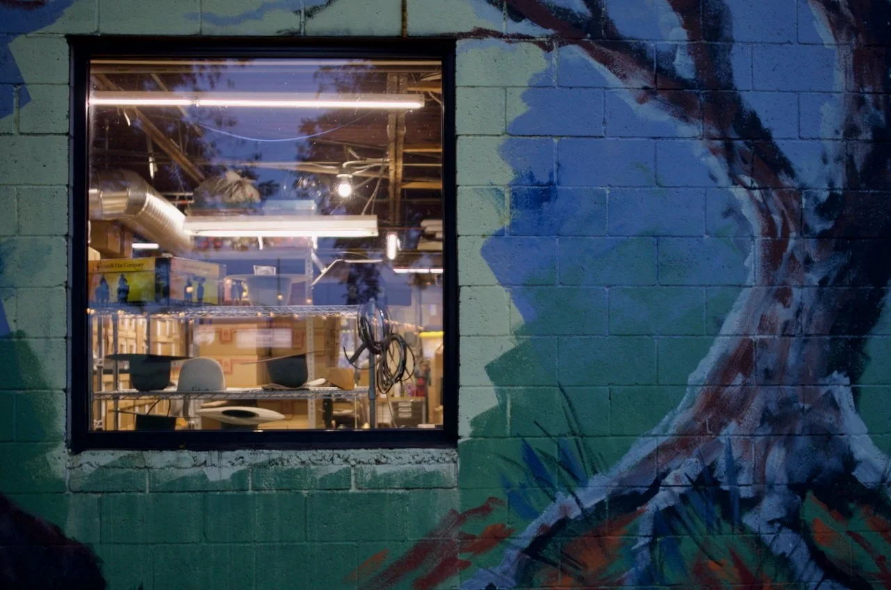 The image shows a window on a brick wall, revealing an interior space that appears to be a workshop or studio with shelves, chairs, and various equipment. The brick wall outside features a colorful mural of a profile of a person's face and hair paint