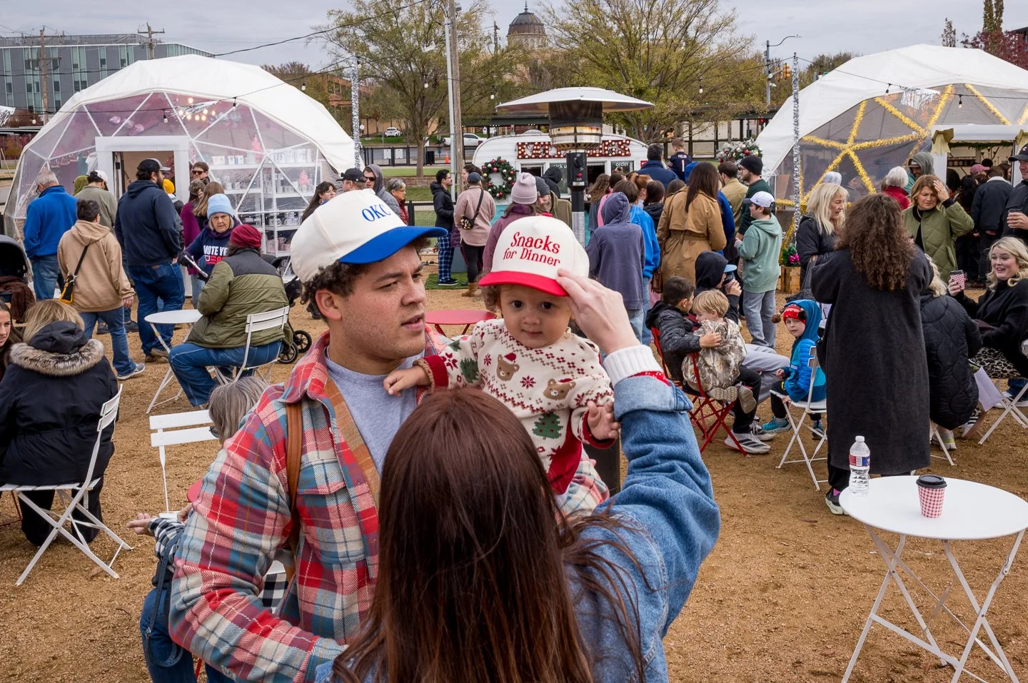 A festive outdoor event with numerous people gathered around, some sitting and some standing, under large white geodesic dome structures, with a food truck in the background and holiday decorations. In the foreground, a man holds a toddler wearing a 