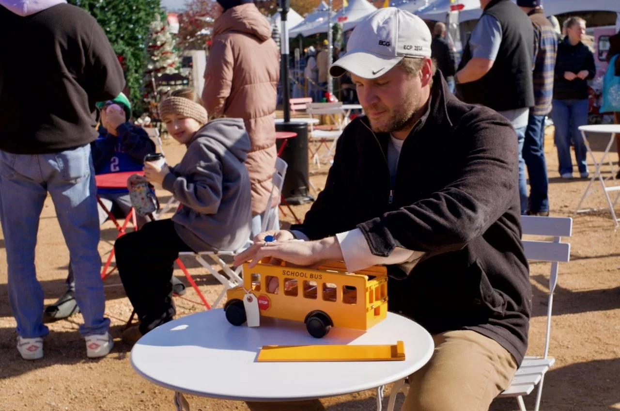 A man assembling a yellow toy school bus model at an outdoor event, with children and other people in the background on a sunny day.