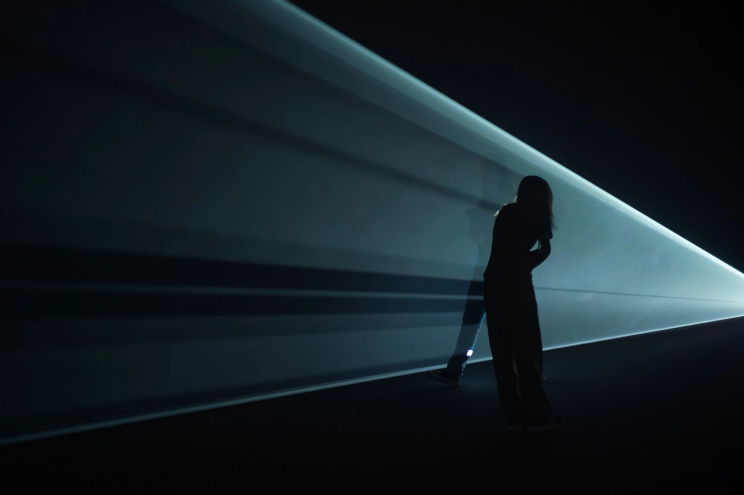 Silhouette of a woman standing near a wall with a bright, linear light source casting soft rays across the dark space.