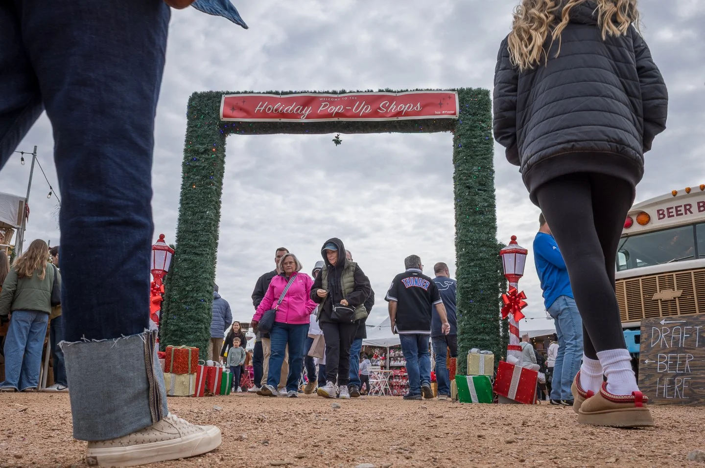 Crowd of people walking under a holiday pop-up shop archway decorated with greenery and lights at an outdoor market, with festive gift boxes at the base of the arch and a food truck in the background.