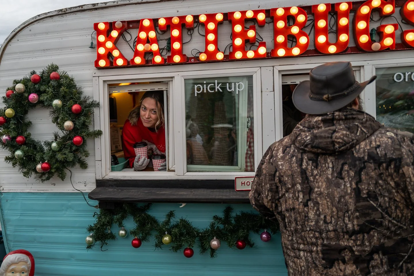 A woman in a festive red sweater is inside a food truck, smiling and holding a hot beverage, as she hands it to a man in camo jacket and cowboy hat outside the truck. The truck is decorated with Christmas wreaths and ornaments, and has large illumina