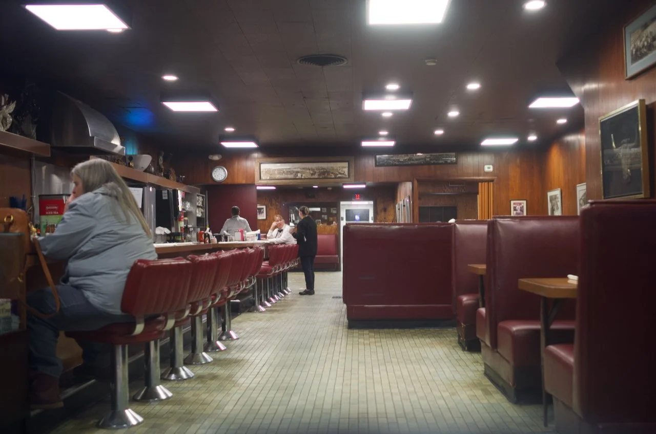 Interior of a retro-style diner with red booths and barstools, wooden-paneled walls, framed pictures, a counter with people and a staff, and overhead ceiling lights.