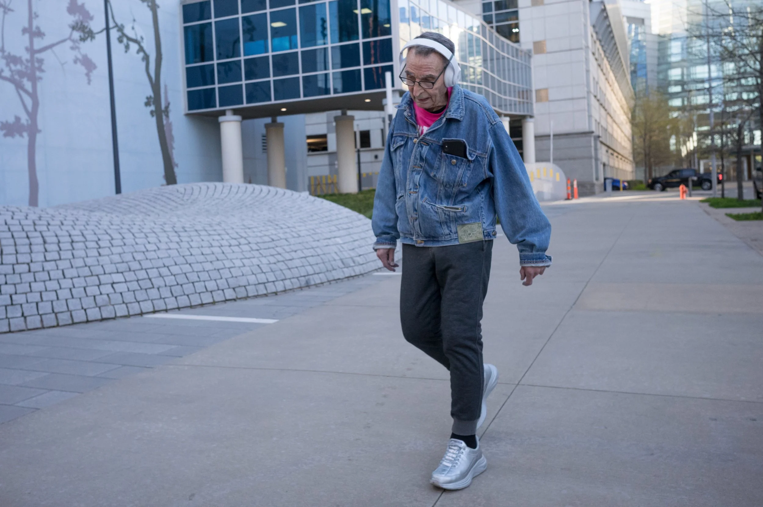An elderly man walking on a city sidewalk wearing headphones, a denim jacket with a pocket, gray sweatpants, and white sneakers, with modern buildings in the background.