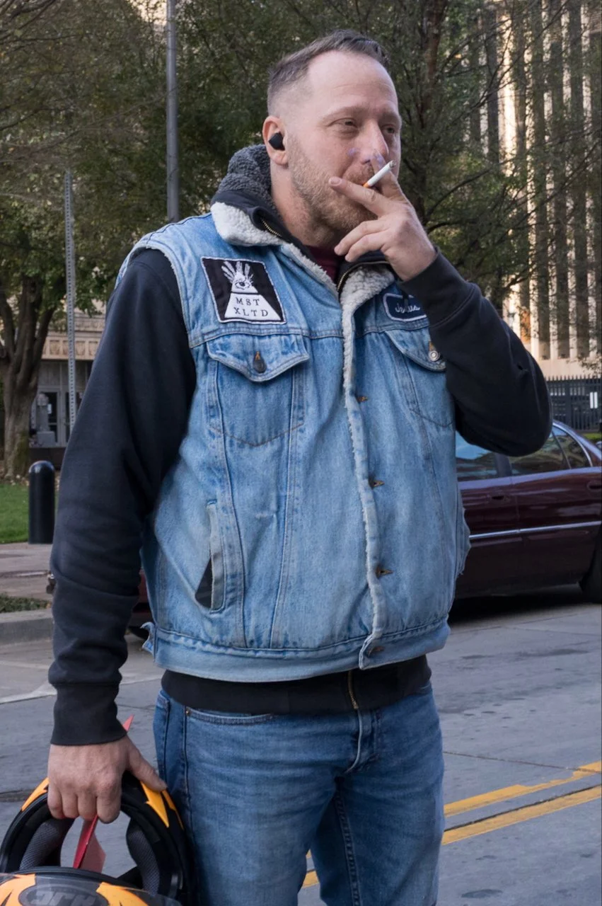 A man with short hair and a beard, wearing a blue denim and black hoodie jacket, standing on a sidewalk, holding a motorcycle helmet in his left hand, with trees and a parked car in the background.