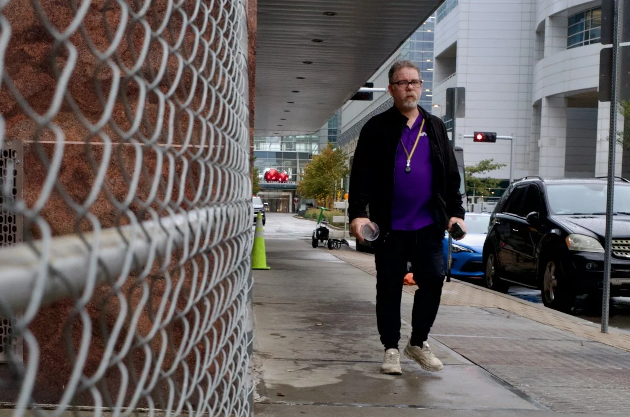 A middle-aged man with glasses and a beard walking on a city sidewalk, carrying water bottles, wearing a purple shirt, black jacket, black pants, and white sneakers. There are cars parked along the street, buildings in the background, and a chain-lin
