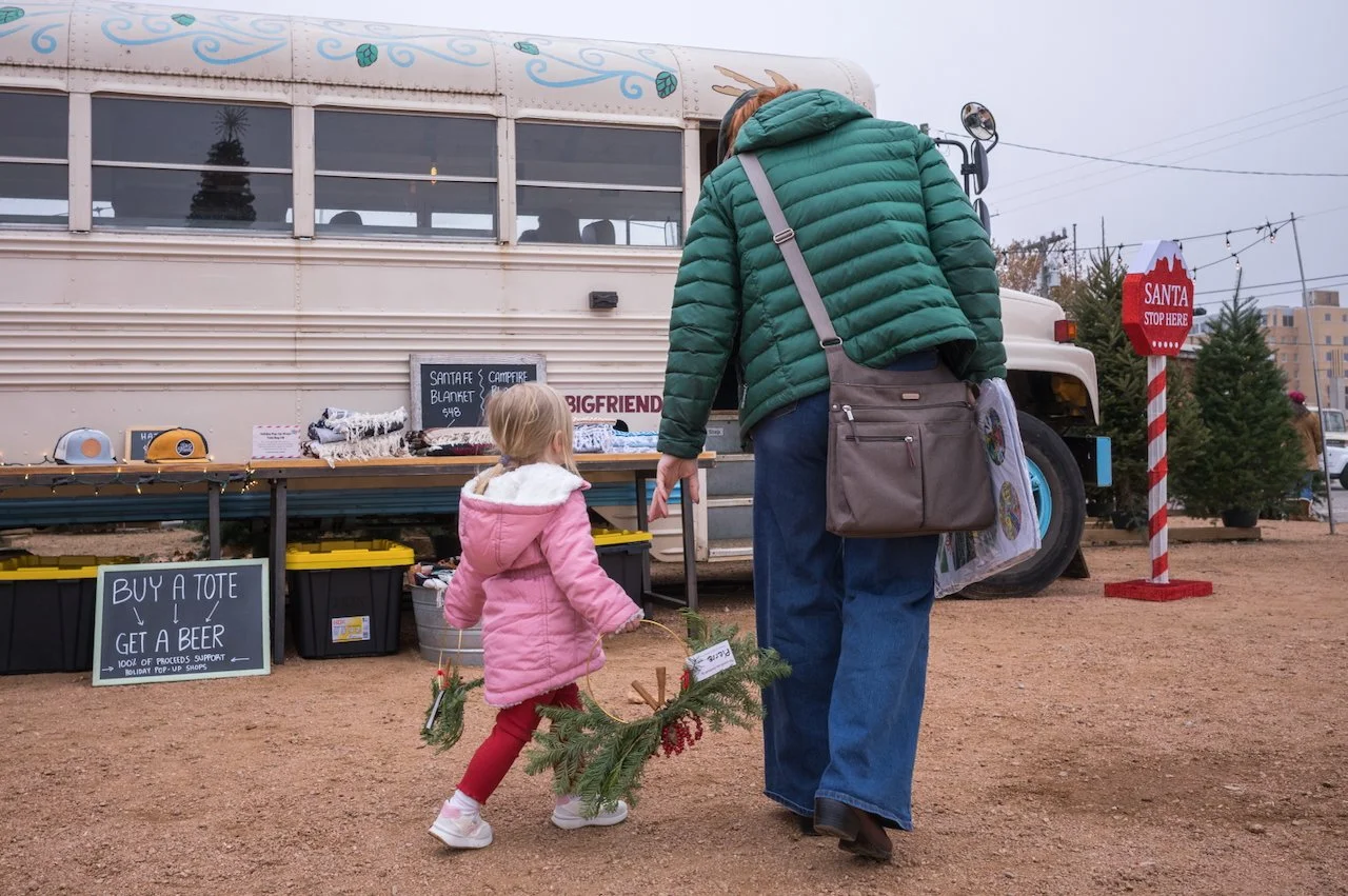 A woman and a young girl walking in front of a Christmas tree lot, the girl holding a festive wreath. The lot has a bus with merchandise for sale, including Santa blankets, and a sign that says "Buy a tote, get a beer." There is a Santa stop sign and