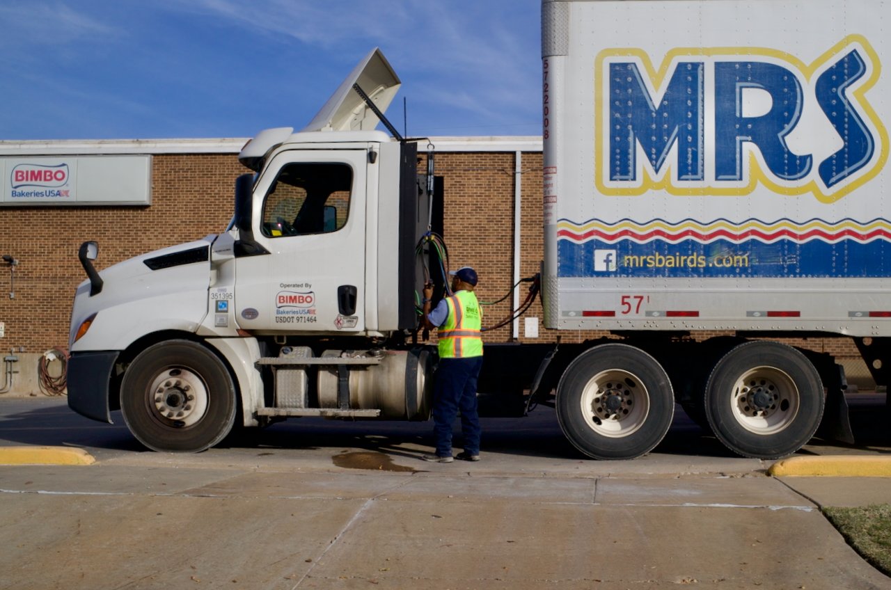 A worker in safety gear standing next to a large delivery truck with a Mr. Baird's logo, parked on a sidewalk in front of a brick building with PPC Bakery's sign, under a clear blue sky.