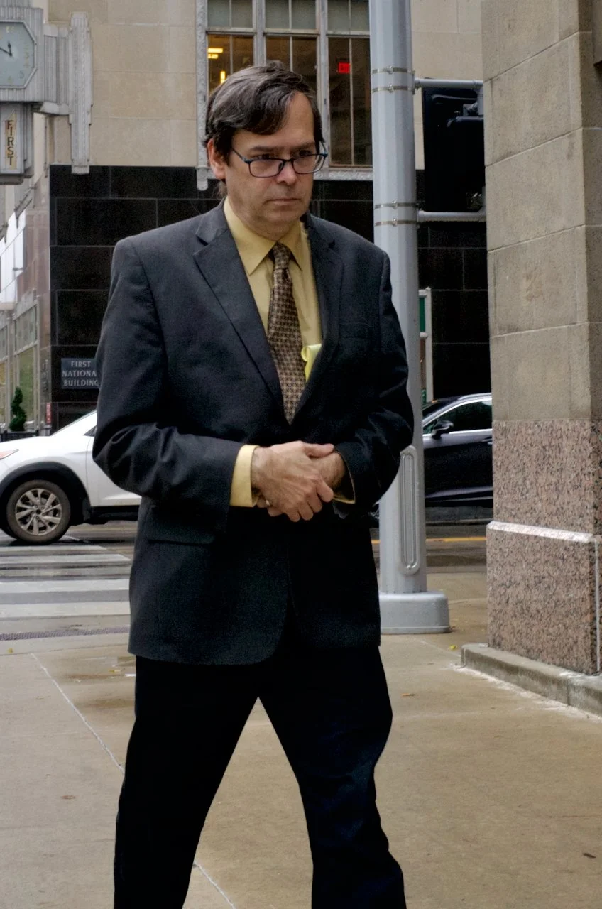 A man in a black suit and glasses walking on a city sidewalk, with office buildings and parked cars in the background.
