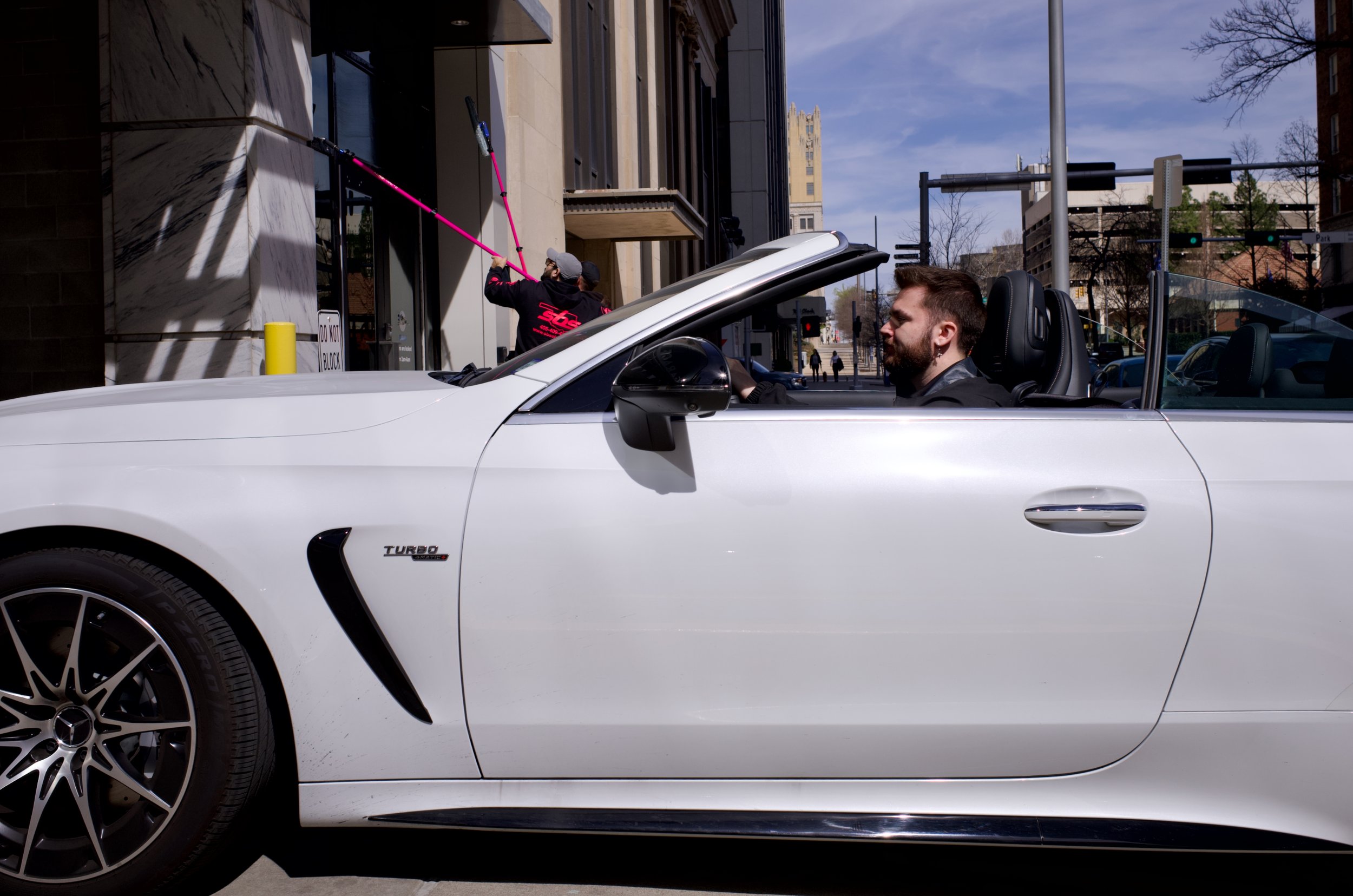 A man with a beard sitting in a white convertible car parked on a city street during the day, with a person in the background using a pink tool on a beige building.