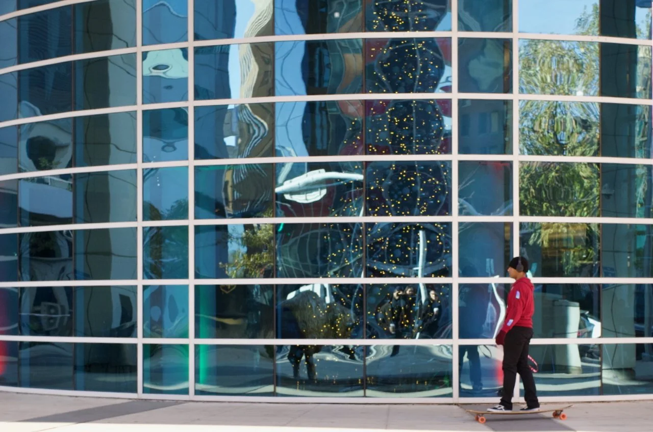 A person in a red hoodie and black pants skateboarding in front of a large glass building with a reflective facade. The reflection shows a spaceship model, trees, and the sky.