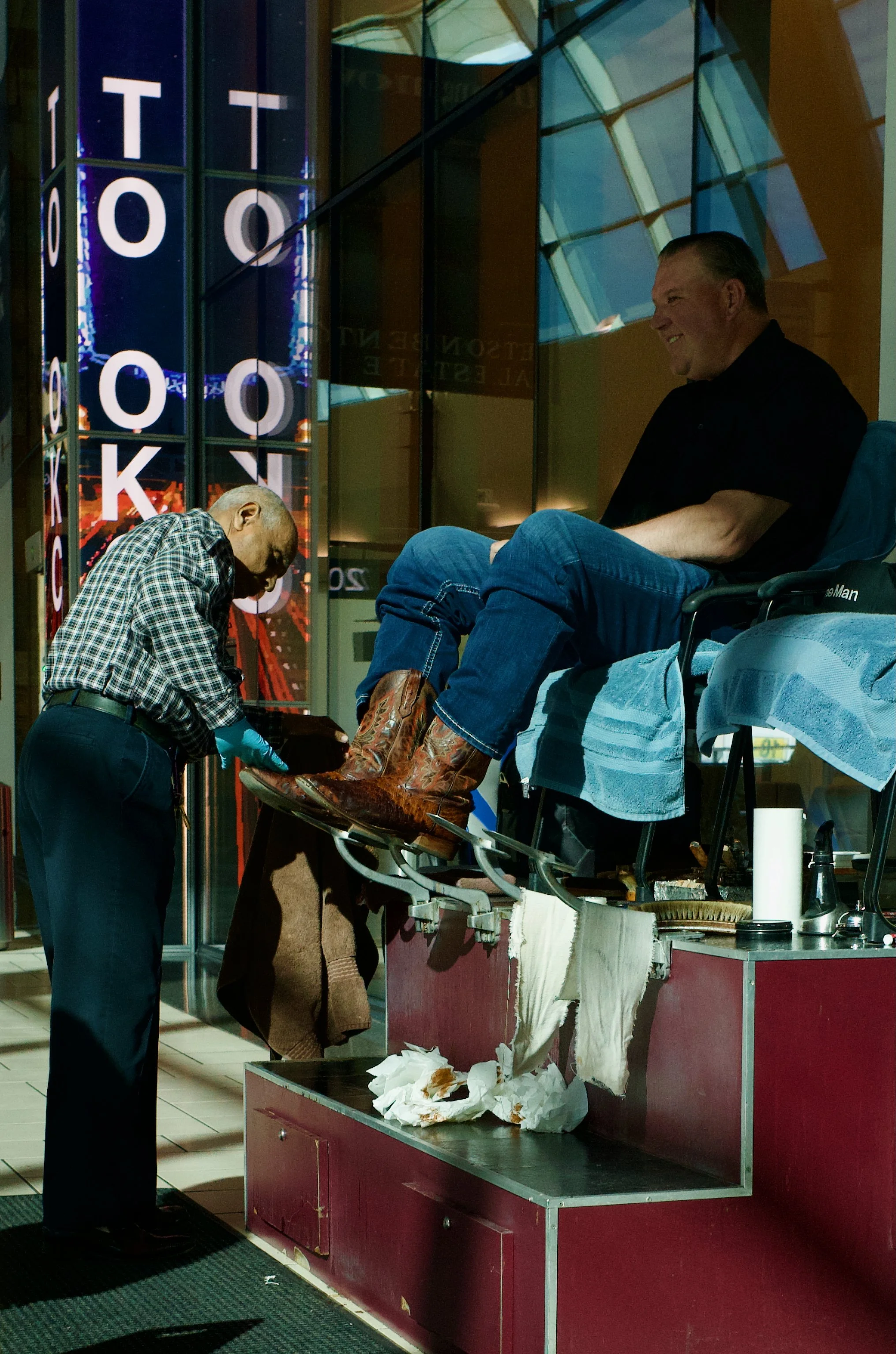 A man in a wheelchair is having cowboy boots fitted by a shoemaker at a shoe repair shop. The man is smiling, wearing jeans and a dark shirt. The shoemaker, an elderly man in a plaid shirt and gloves, is working on the boots. The scene is inside a mo