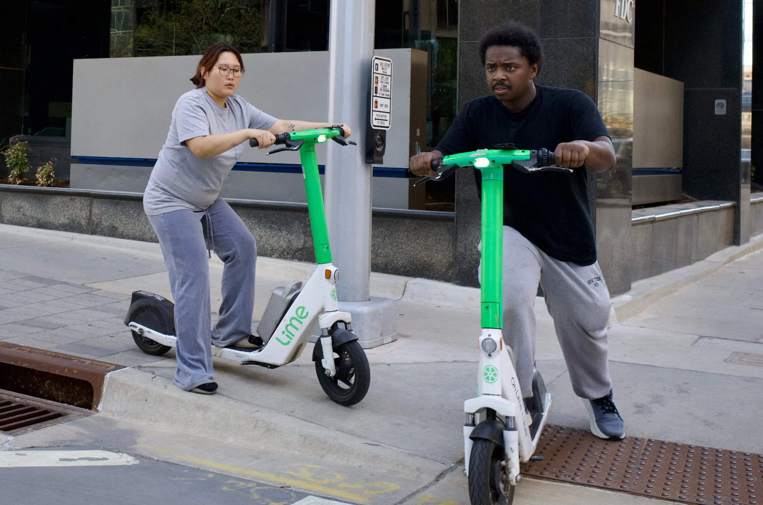 Two people riding Lime electric scooters on a city sidewalk, with a woman in gray shirt and pants on the left and a man in black shirt and gray sweatpants on the right.