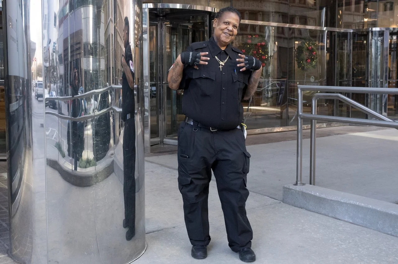 A woman in a police uniform standing outside a building, smiling, making a peace sign with both hands, wearing fingerless gloves and necklaces, with Christmas wreaths visible through the glass doors behind her.