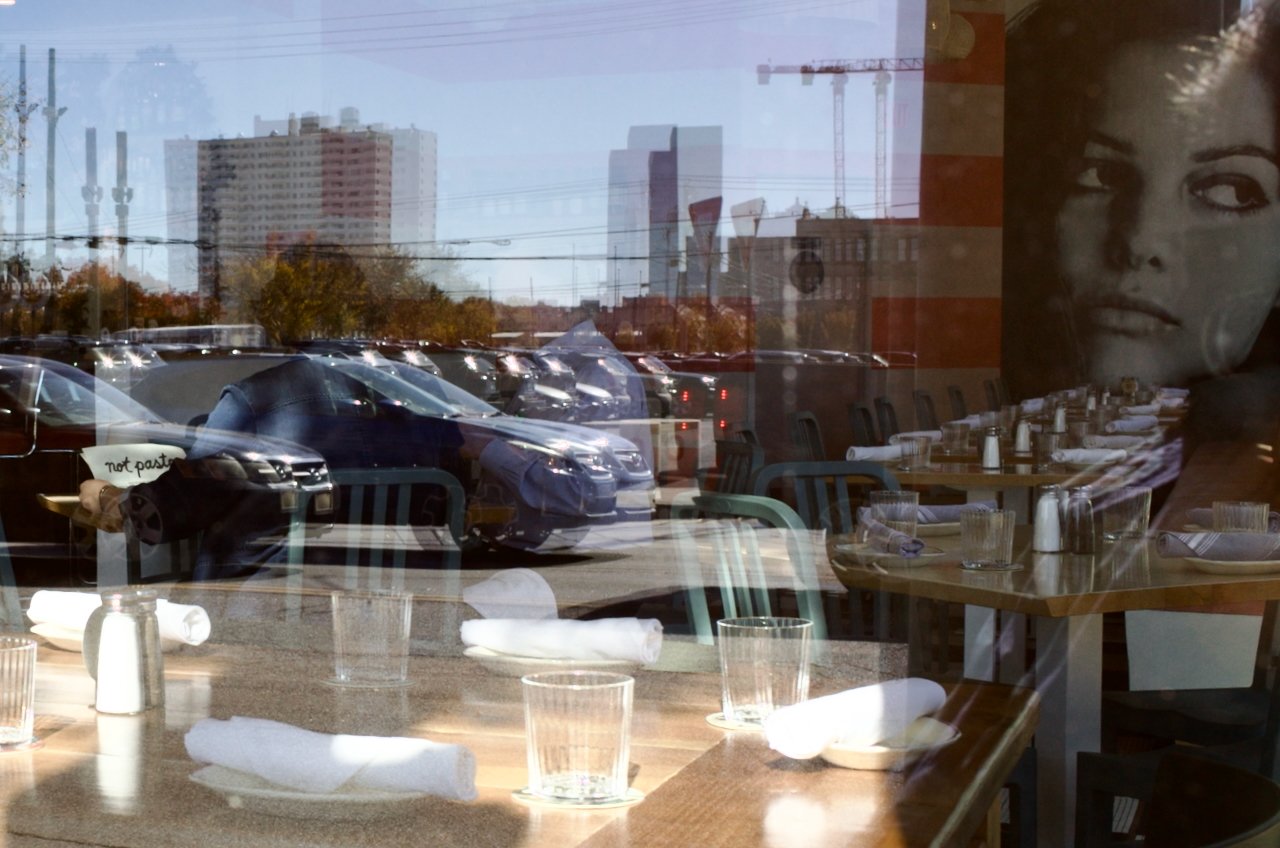Reflections of parked cars and buildings outside through a restaurant window, with a woman's face partially visible on the right side of the window