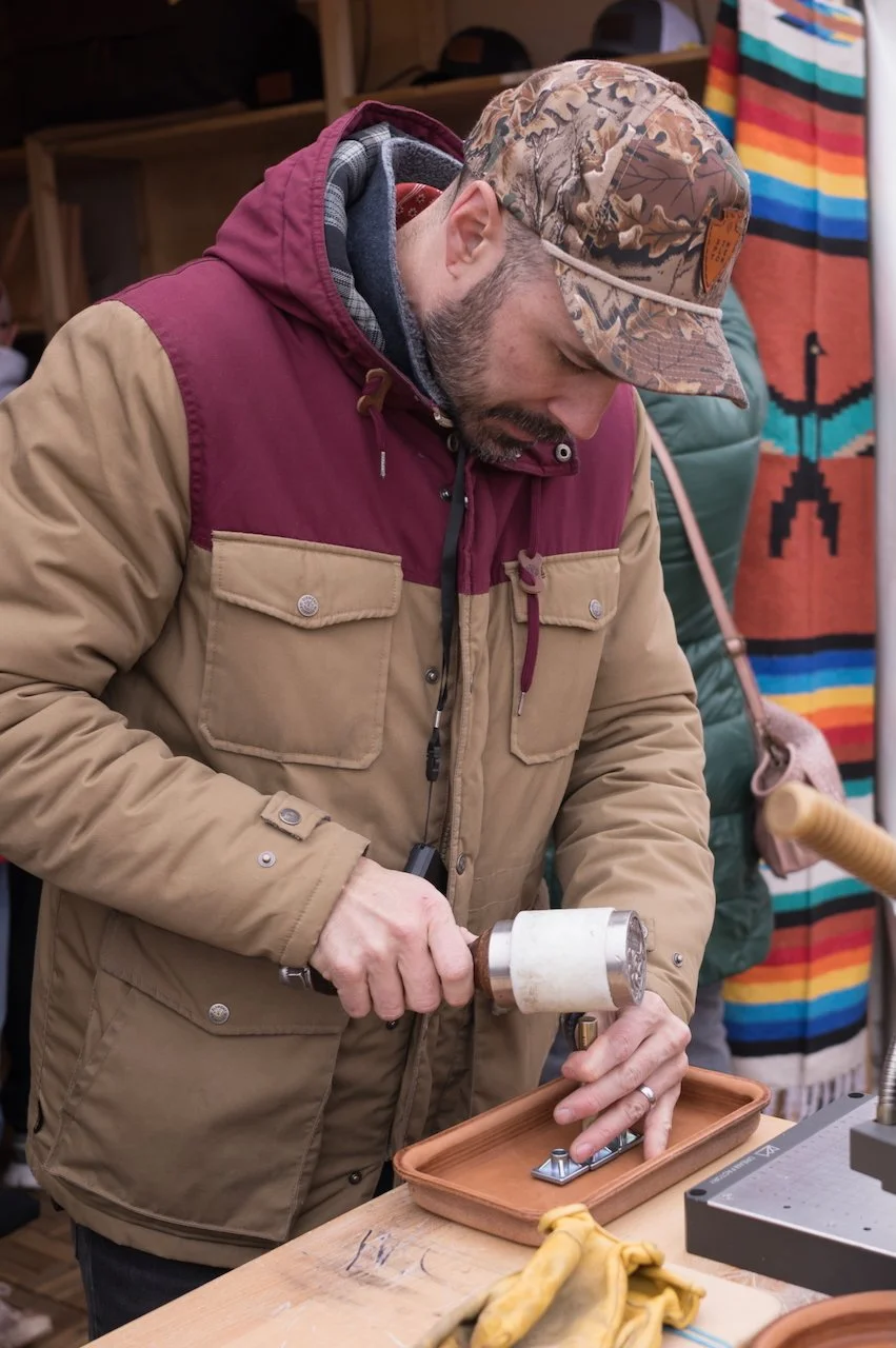 A man wearing a camouflage cap and a beige and maroon jacket is working on a project with a mallet and a metal hinge in a workshop.