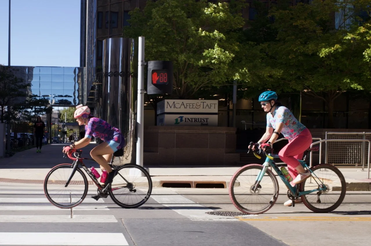 Two women riding bicycles across a city street with a crosswalk. One woman is wearing a pink helmet and colorful athletic clothing, and the other is wearing a blue helmet and floral shirt with pink pants. There are buildings and trees behind them.