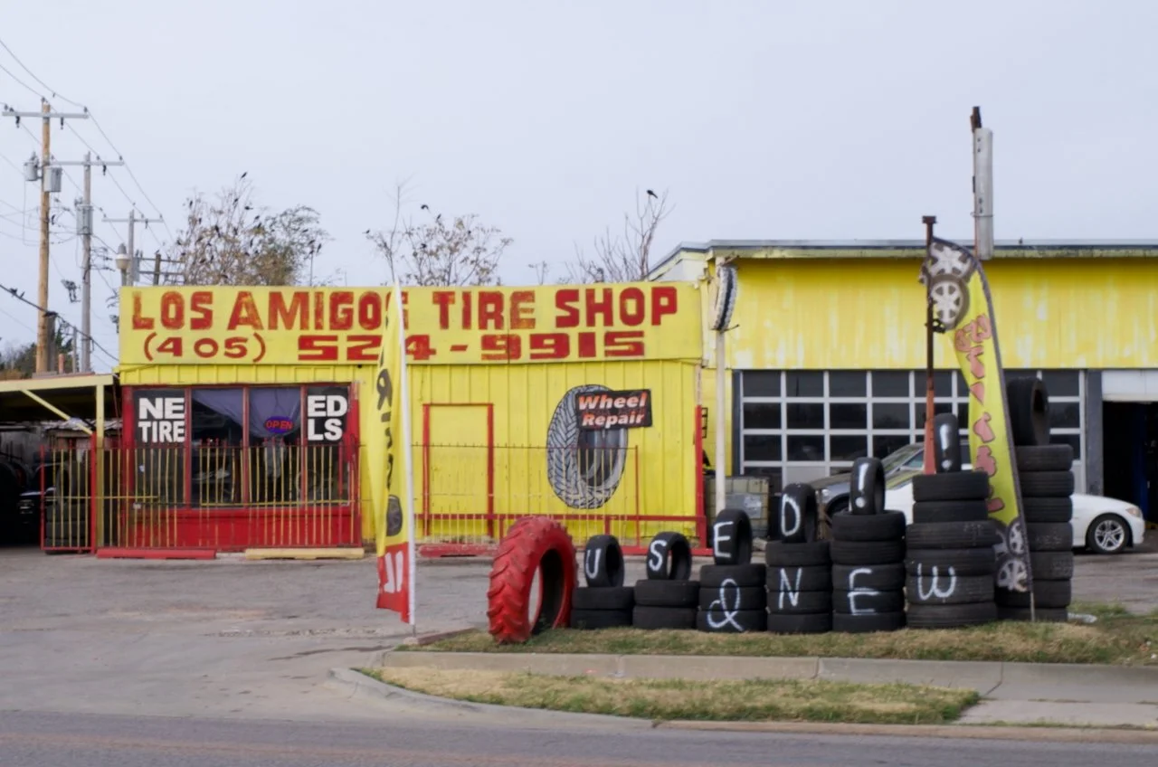 Outside view of Los Amigo Tire Shop painted yellow with red lettering. Several stacked tires with white letters and flags advertising tire services, and a red tire displayed outside. Power lines and trees are in the background.