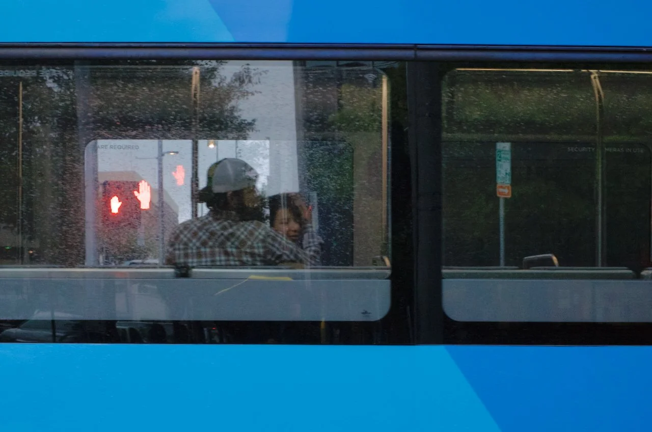 Two people sitting inside a bus through a window, one wearing a hat and checkered shirt, the other woman looking towards the camera. The bus window has raindrops on it, and a red hand symbol is visible on a nearby sign.