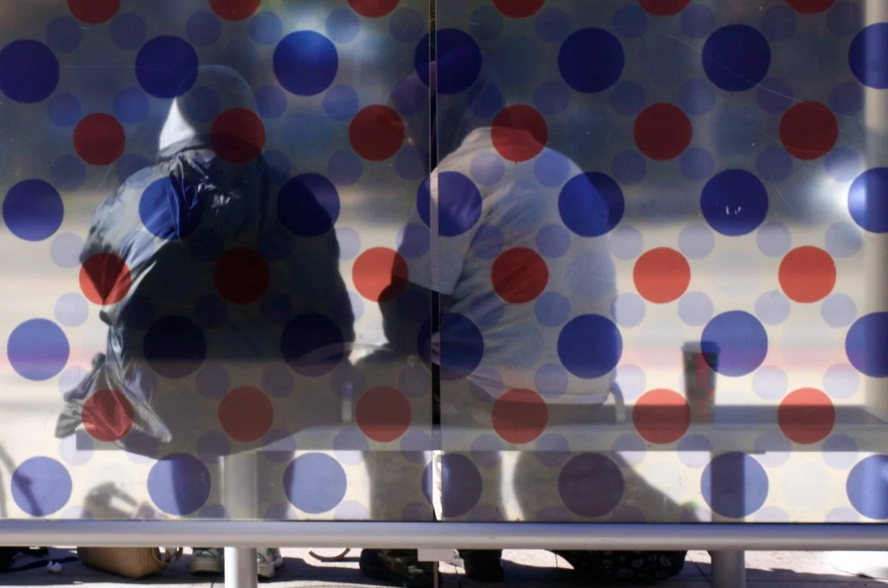 Two individuals sitting at a table behind a glass window design with red and blue dots, one with a hoodie and the other with a white shirt, along with a beverage in a cup on the table.