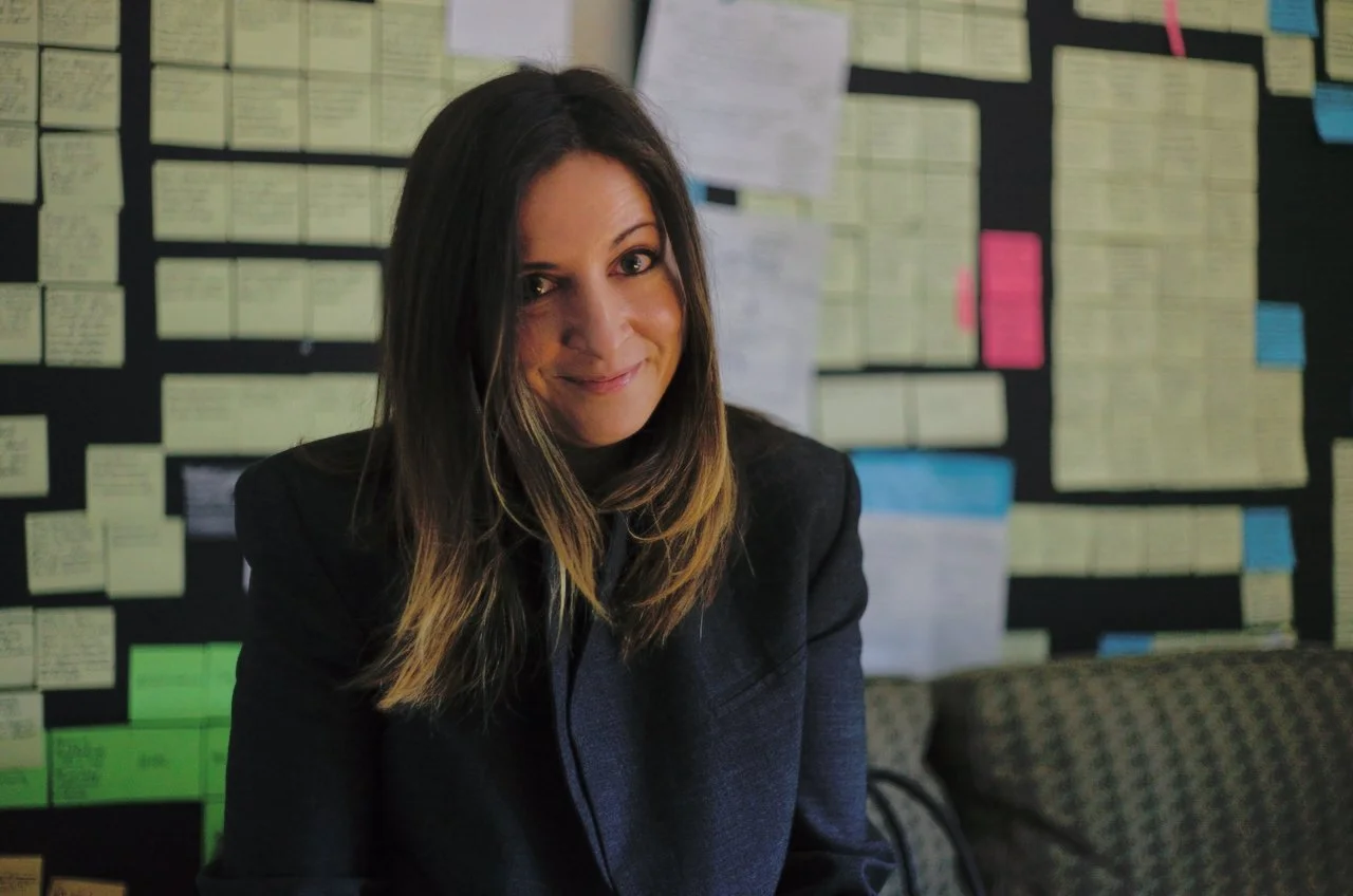 A woman with shoulder-length dark hair, smiling and looking at the camera, sitting in front of a wall covered with colorful sticky notes and papers.
