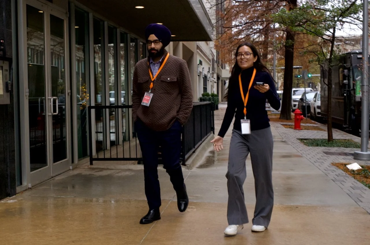 Two people, a man with a turban and a woman with glasses, walking and talking on a city sidewalk in autumn.