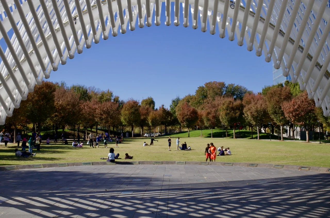 Park scene with people sitting and walking under a clear blue sky, framed by white architectural structure and colorful trees.