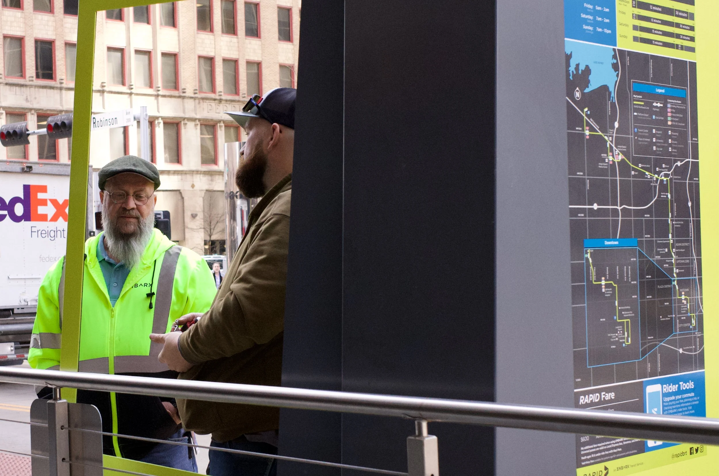 Two men are standing near a information board or map at an urban transit station; one is wearing a bright yellow safety jacket and a gray flat cap, and the other is dressed in a brown jacket and a baseball cap, both are engaged in conversation.
