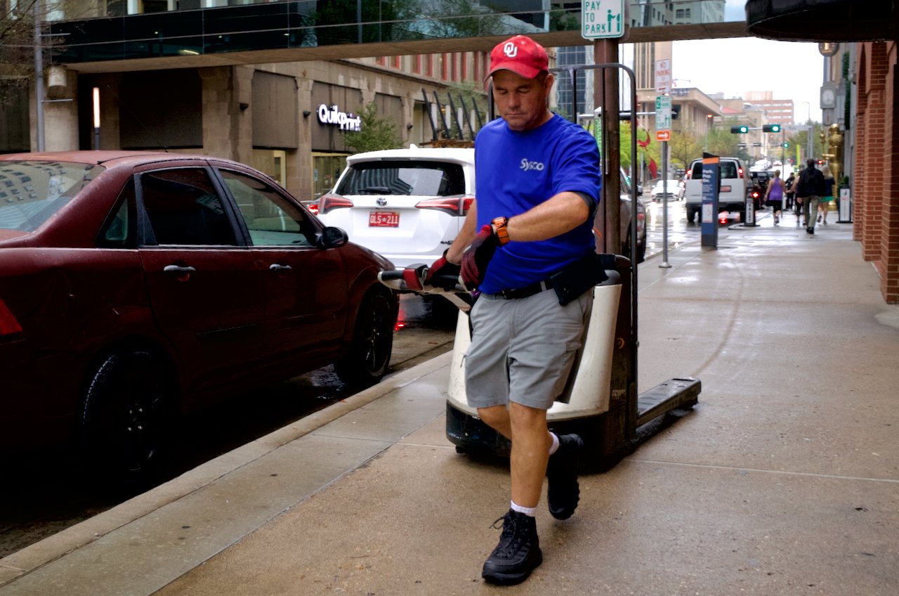 A delivery person wearing a blue shirt, gray shorts, and a red cap walking on a city sidewalk pulling a hand truck or dolly with a package on it, next to parked cars and buildings.