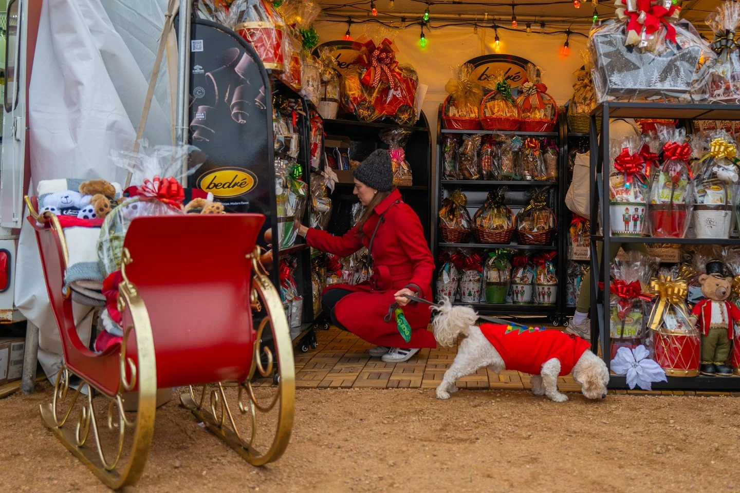 A woman in a red coat and gray beanie is shopping at a holiday market stall, while a small dog in a red sweater stands nearby. The stall is decorated with Christmas items and gift baskets.