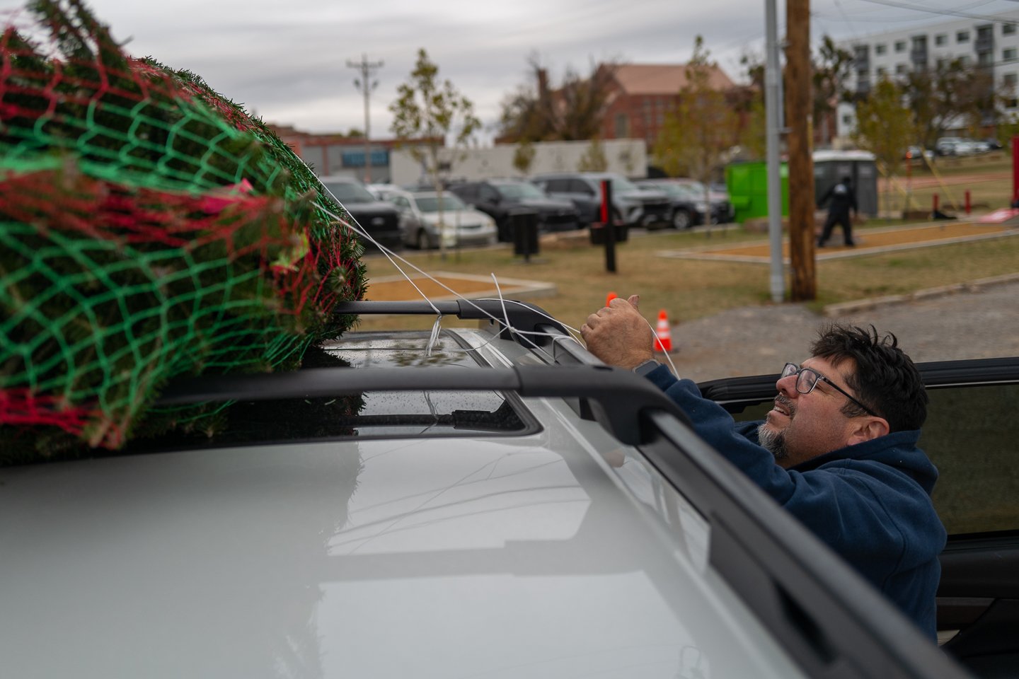 A man with glasses and a beard is placing a Christmas tree on top of his car, tying it with strings, in a parking lot on a cloudy day.