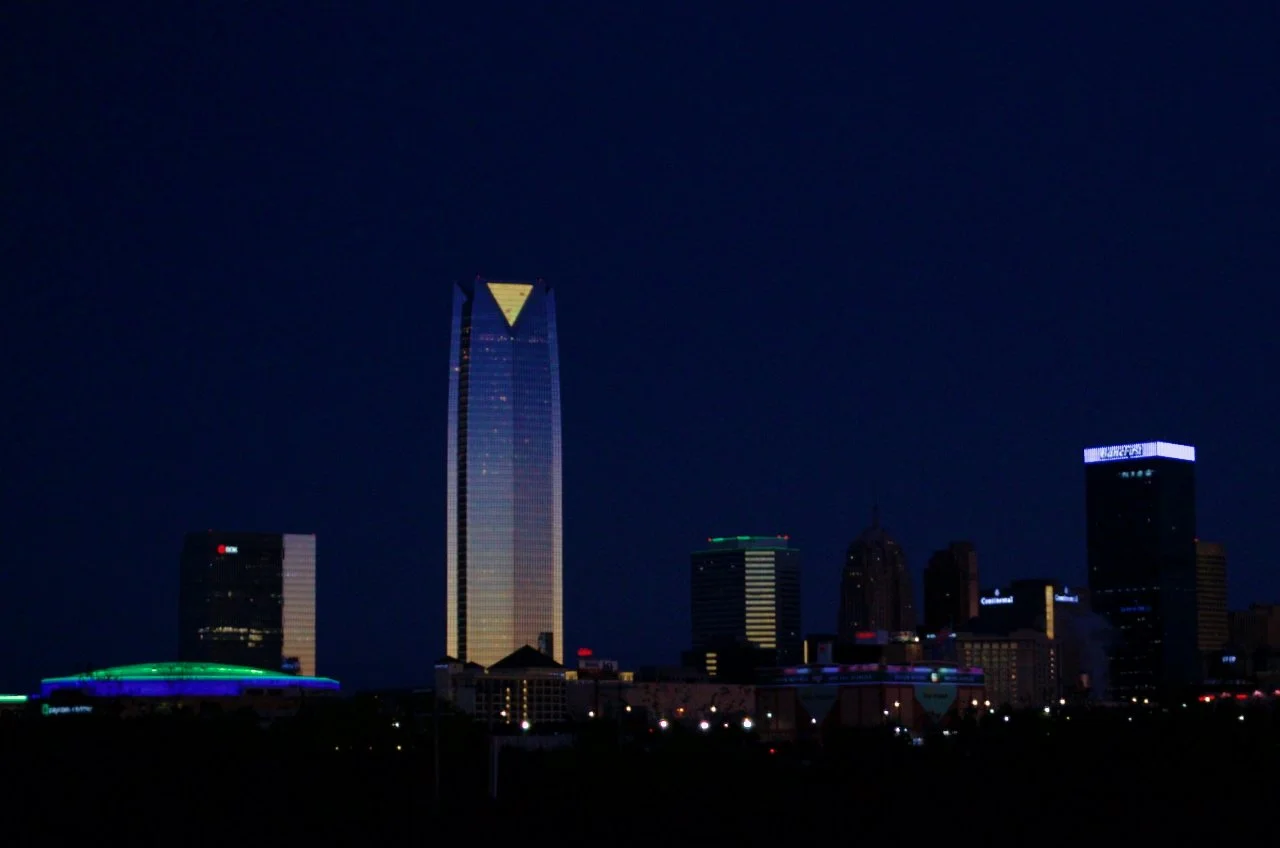 Nighttime cityscape with illuminated skyscrapers, including a tall building with a triangular top, in an urban area.